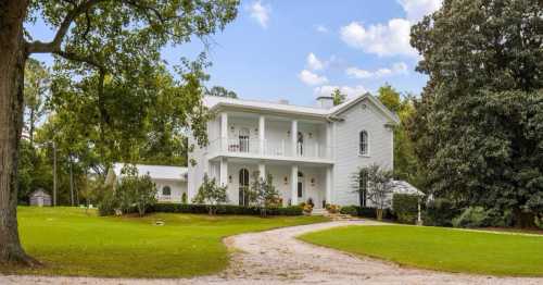 A charming white house with a porch, surrounded by green grass and trees, set against a blue sky with fluffy clouds.