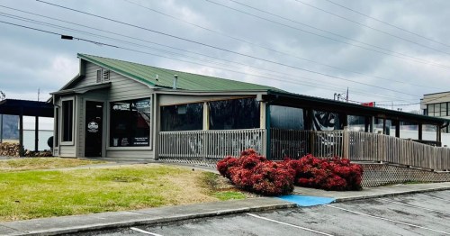 A single-story building with a green roof, large windows, and a deck, surrounded by grass and red bushes.
