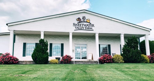 Exterior of Sweetwater Valley Farm building with green grass and colorful flower beds under a cloudy sky.