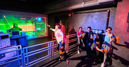 A group of six people play a laser tag game in a colorful, futuristic setting with neon lights and a digital screen.