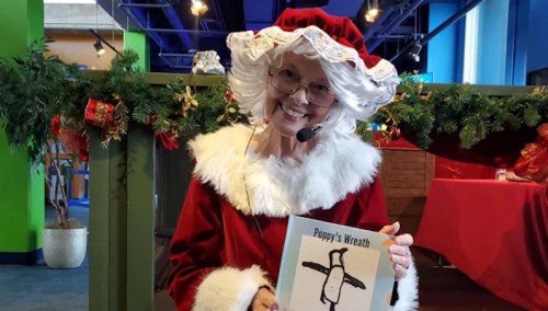 A smiling woman in a Santa costume holds a children's book titled "Poppy's Wreath" in a festive setting.