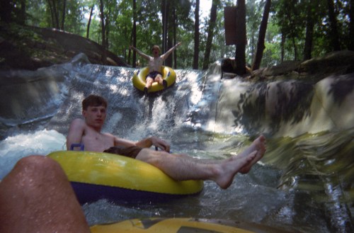 Two people on inner tubes slide down a water slide surrounded by trees, enjoying a summer day.