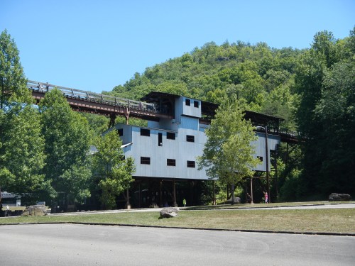 A modern building on stilts surrounded by trees, with a train track running above it against a green hillside.