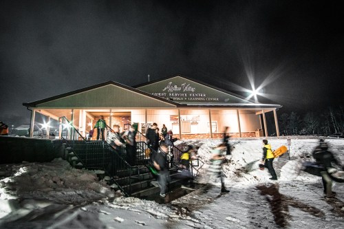 A group of people exits a building at night, surrounded by snow, with bright lights illuminating the scene.