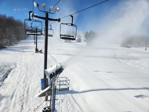 Snowmaking equipment sprays snow on a ski slope, with a chairlift and clear blue sky in the background.