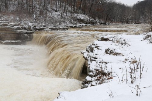 A snowy landscape featuring a waterfall cascading over rocky cliffs into a river below.