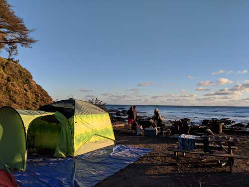 A green tent by the shore, with people preparing for camping, and a scenic view of the ocean and sky.