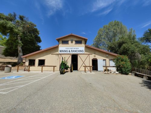 A rustic building with a sign reading "Mining & Ranching," featuring a green tractor in front and trees in the background.