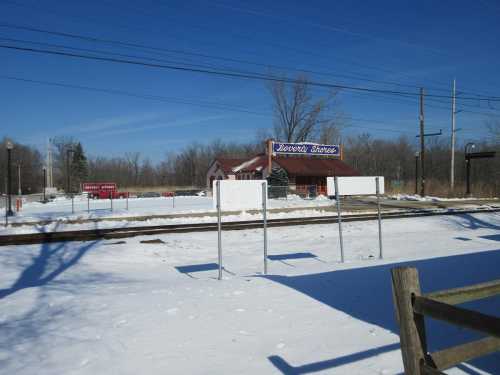 A snowy landscape with a train station in the background, featuring a red building and empty signposts.