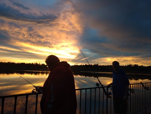 Two people fish by a calm lake at sunset, with vibrant clouds reflecting in the water.