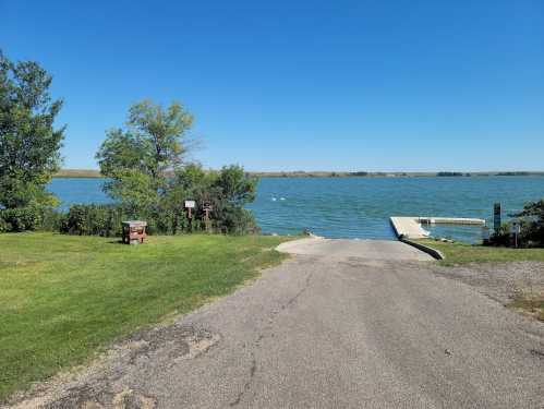 A serene lake view with a grassy area, a bench, and a boat ramp under a clear blue sky.