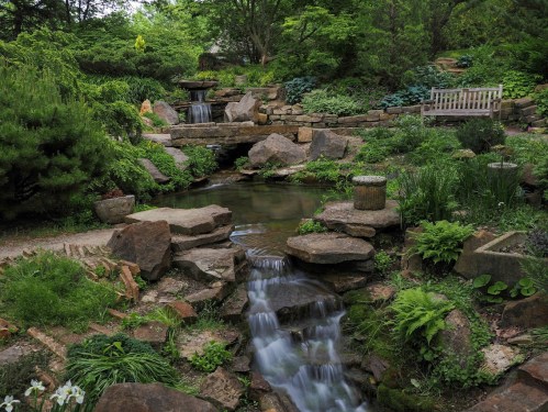 A serene garden scene featuring a flowing stream, rocks, lush greenery, and a wooden bench amidst the natural landscape.