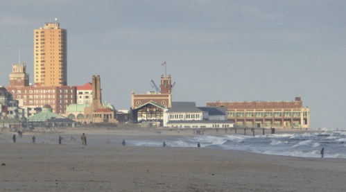 A beach scene with people walking along the shore, featuring buildings and a boardwalk in the background.