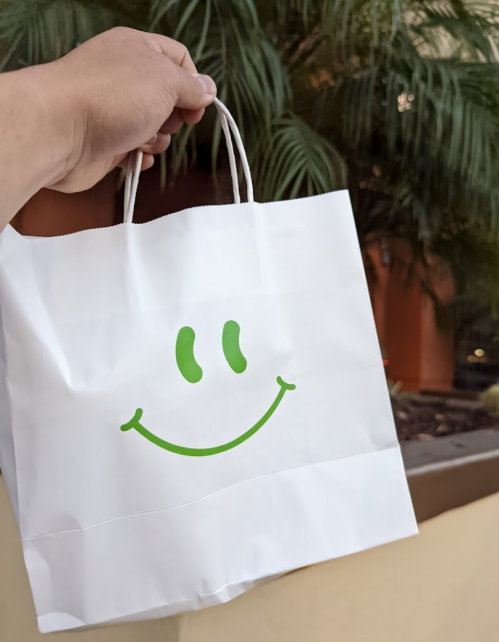 A hand holds a white shopping bag featuring a green smiley face against a backdrop of palm leaves.