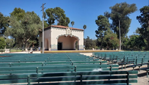 An empty outdoor amphitheater with green benches and a decorative stage surrounded by trees under a clear blue sky.