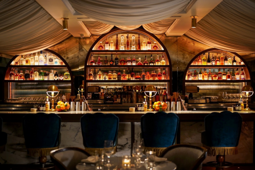 A stylish bar with illuminated shelves of liquor, elegant seating, and soft drapery overhead.