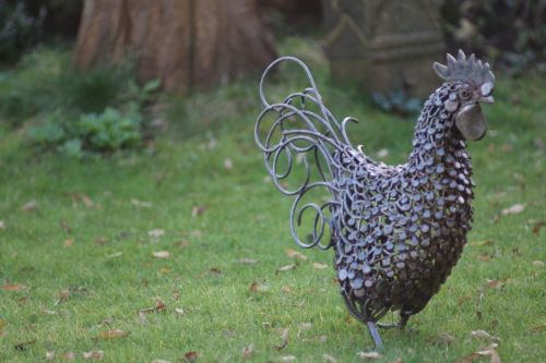 A decorative metal sculpture of a rooster stands on green grass, surrounded by foliage.