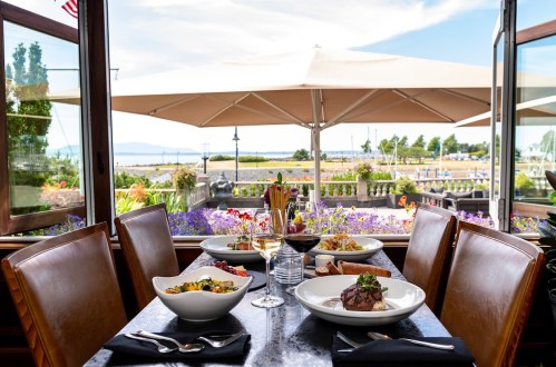 A beautifully set table with gourmet dishes and wine, overlooking a scenic outdoor view with flowers and a waterfront.