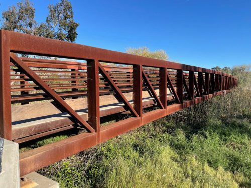 A rust-colored metal bridge spans a grassy area under a clear blue sky.