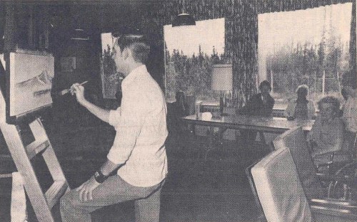 A man paints on an easel while a group of people sits at a table in a room with large windows overlooking trees.