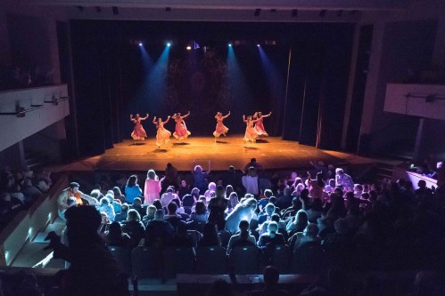 A stage performance with dancers in colorful costumes, illuminated by stage lights, while an audience watches from below.