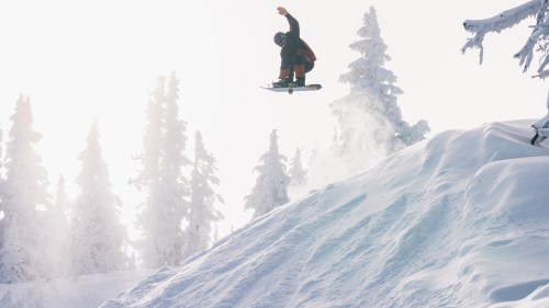 A snowboarder jumps off a snowy slope, surrounded by tall, snow-covered trees and bright sunlight.