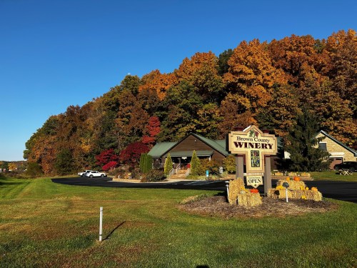 A winery surrounded by vibrant autumn foliage and a clear blue sky, with a sign indicating it's open for visitors.