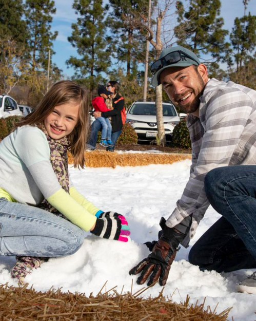 A man and a girl smile while playing in the snow, surrounded by trees and people in the background.