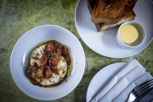 A bowl of poached eggs with tomatoes and herbs, accompanied by toasted bread and a small dish of butter.