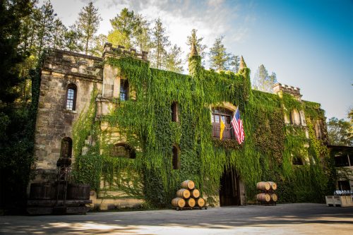 A historic stone building covered in greenery, with an American flag and wine barrels in front, set against a blue sky.