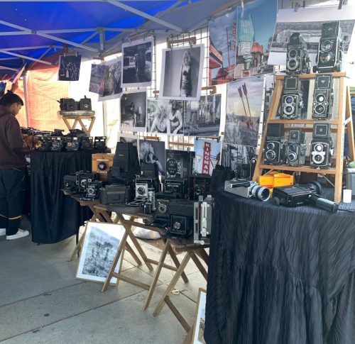 A market stall displaying vintage cameras and black-and-white photographs, with a person browsing in the background.