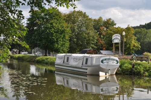 A boat named "St. Hellen's IV" moored on a calm canal, surrounded by trees and a sign in the background.