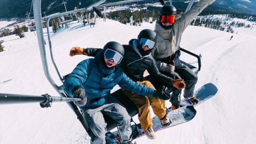 Three friends on a ski lift, smiling and posing for a selfie, surrounded by snowy mountains and a clear blue sky.