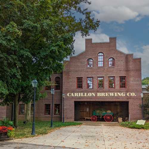 A brick building with large windows and the sign "Carillon Brewing Co." surrounded by trees and a vintage wagon.