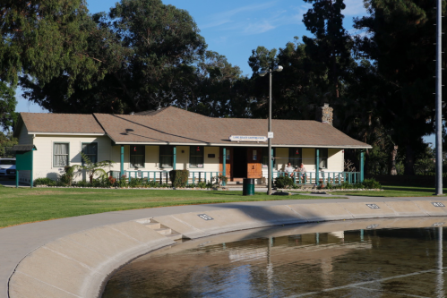 A single-story building with a brown roof, surrounded by greenery and a pond, under a clear blue sky.