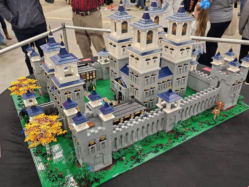A detailed LEGO castle model with towers, walls, and surrounding trees, displayed at an event.