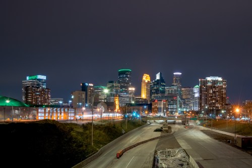 Minneapolis skyline at night, featuring illuminated buildings and a clear sky, with a highway in the foreground.