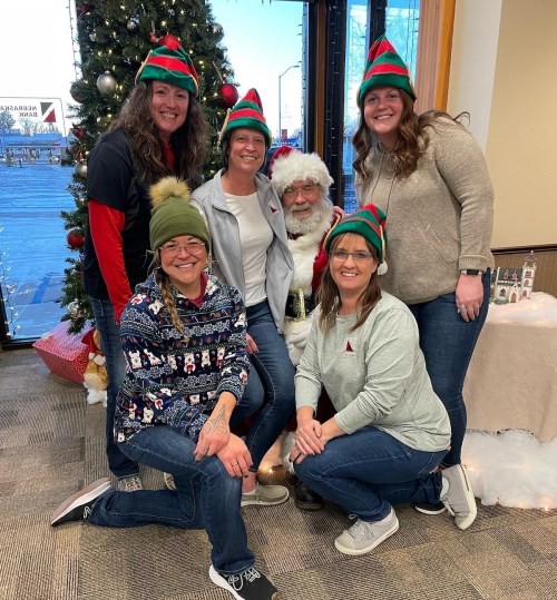 A group of six people, wearing festive hats, pose with Santa in front of a decorated Christmas tree.
