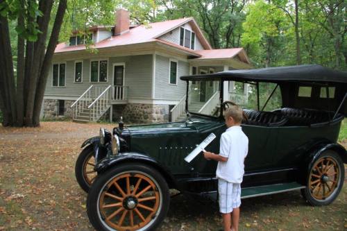 A boy stands next to a vintage car in front of a large house surrounded by trees.
