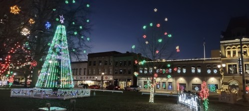 A festive park scene at night, featuring a brightly lit Christmas tree and colorful holiday decorations on buildings.