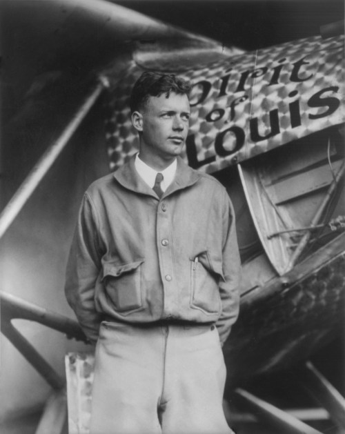 A man stands beside the aircraft "Spirit of St. Louis," wearing a jacket and looking confidently at the camera.