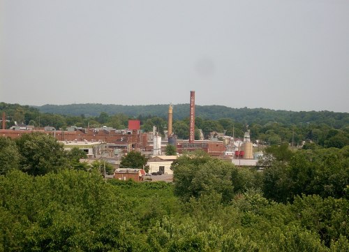 A view of an industrial area with factories and smokestacks, surrounded by green trees and hills in the background.
