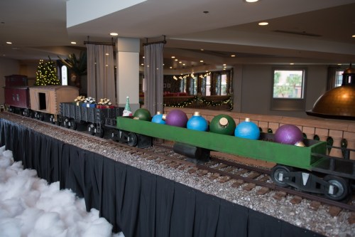 A decorative train display with colorful ornaments on a green flatcar, set indoors with festive decor in the background.