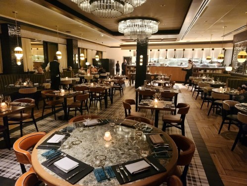 Elegant restaurant interior with round tables, soft lighting, and a stylish bar area in the background.