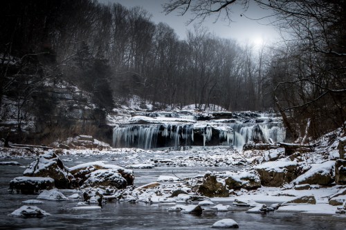 A serene winter scene featuring a frozen waterfall surrounded by snow-covered rocks and trees under a cloudy sky.