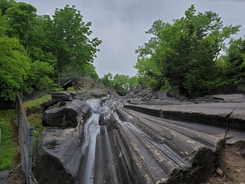 A rocky landscape with smooth, sloped surfaces, surrounded by lush green trees under a cloudy sky.
