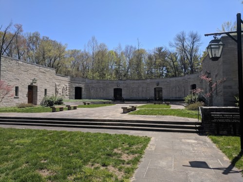 A serene courtyard with stone walls, benches, and greenery, surrounded by trees under a clear blue sky.