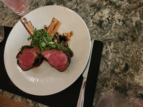 A plate of medium-rare lamb with herb crust, garnished with microgreens and served with vegetables and bone marrow.