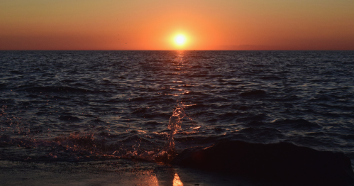 A serene sunset over the ocean, with waves gently splashing against a rocky shore.