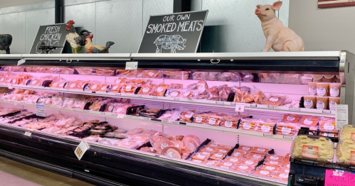A meat display case featuring various smoked meats and fresh chicken, with a decorative pig and chickens above.